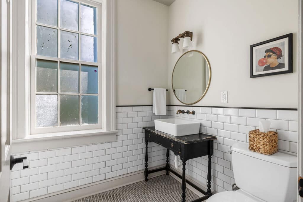 Small vintage-style bathroom with white subway tile, black wooden vanity, square white vessel sink, and round gold mirror under a frosted window.