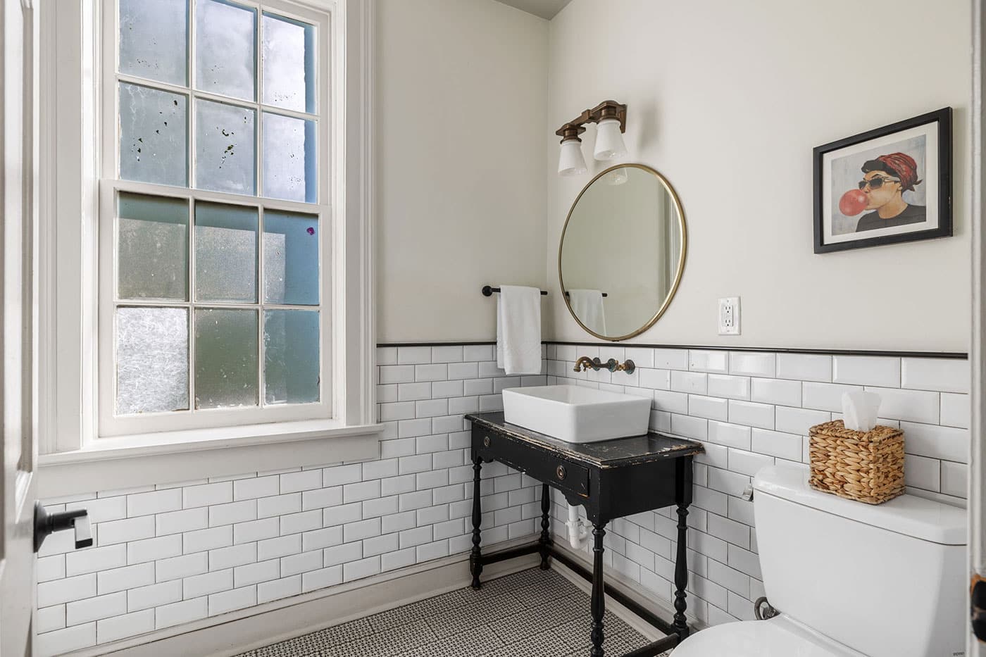 Small vintage-style bathroom with white subway tile, black wooden vanity, square white vessel sink, and round gold mirror under a frosted window.