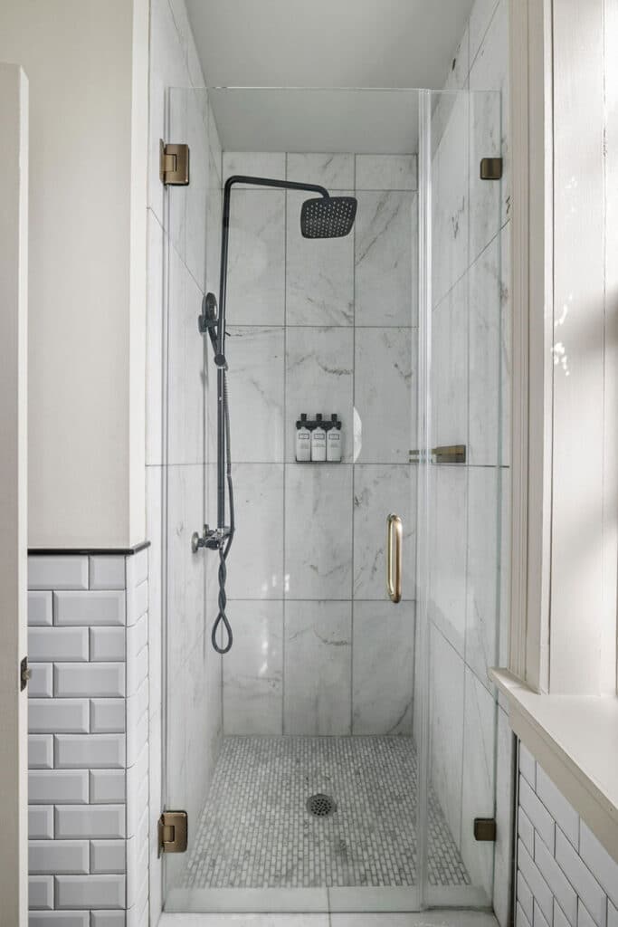 Glass-walled shower with a large black rainfall head, handheld shower, and a small shelf of toiletries on marble-tiled wall, pebble floor mat