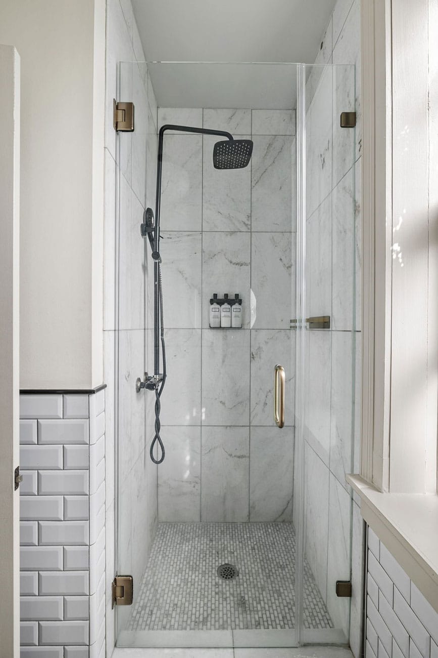 Glass-walled shower with a large black rainfall head, handheld shower, and a small shelf of toiletries on marble-tiled wall, pebble floor mat