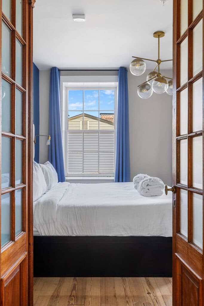 Bright bedroom with white bedding and blue curtains, viewed through an open wooden door, towels rolled on the bed.