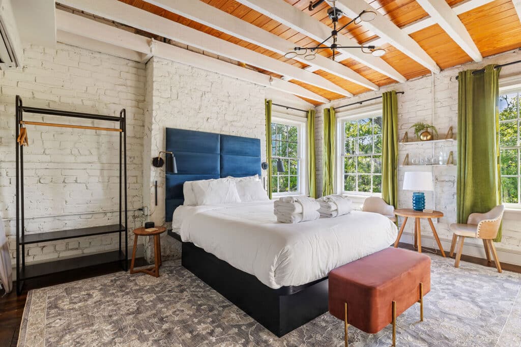 Bright bedroom with white brick walls, exposed wooden beams, blue upholstered headboard, and white bedding by large windows with green curtains.