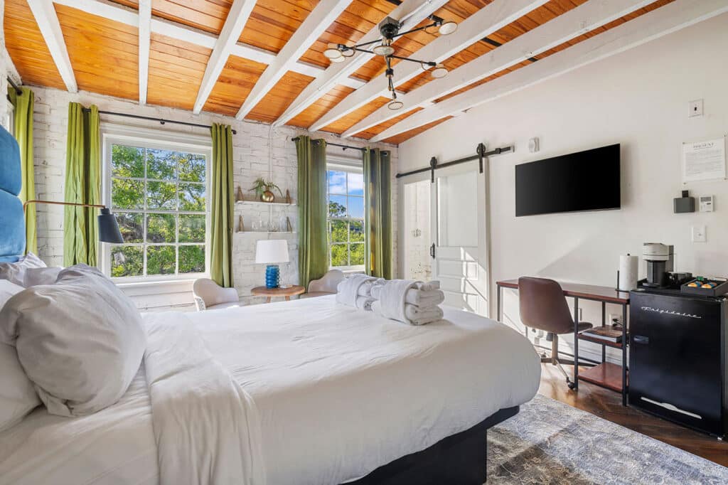 Bright bedroom with white bedding and rolled towels on the bed, exposed white-painted beams, two windows with green curtains, a small seating area, wall-mounted TV, and a desk with a minibar/coffee station.