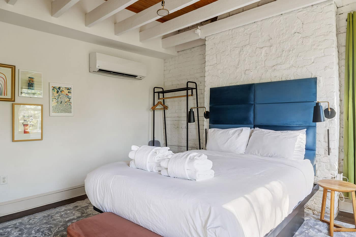 King-size bed with white bedding and a blue upholstered headboard against a white brick wall, with rolled towels on the duvet.