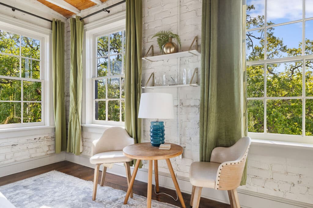 Bright reading nook with white brick wall, three windows, green curtains, and a round wooden table between two beige chairs.