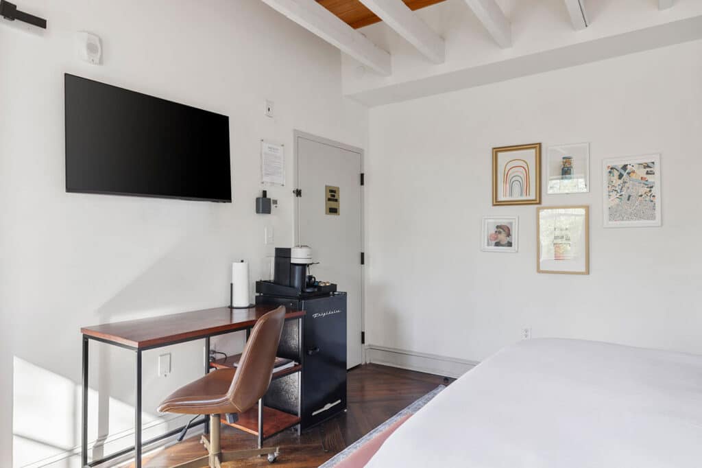 Desk area in a bright bedroom: wall-mounted TV, wooden desk, brown chair, coffee station, and mini-fridge under a small cabinet with framed art on the wall to the right.