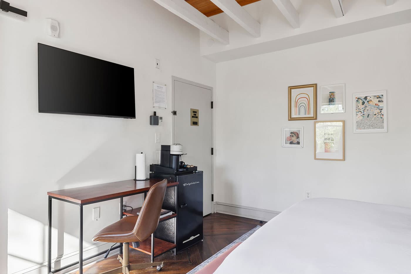Desk area in a bright bedroom: wall-mounted TV, wooden desk, brown chair, coffee station, and mini-fridge under a small cabinet with framed art on the wall to the right.