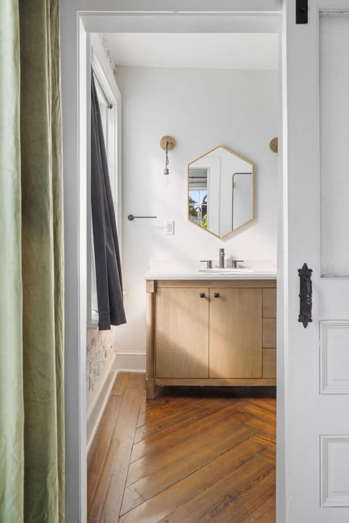 View into a small bathroom with a wooden vanity, hexagonal mirror, and warm wood floor