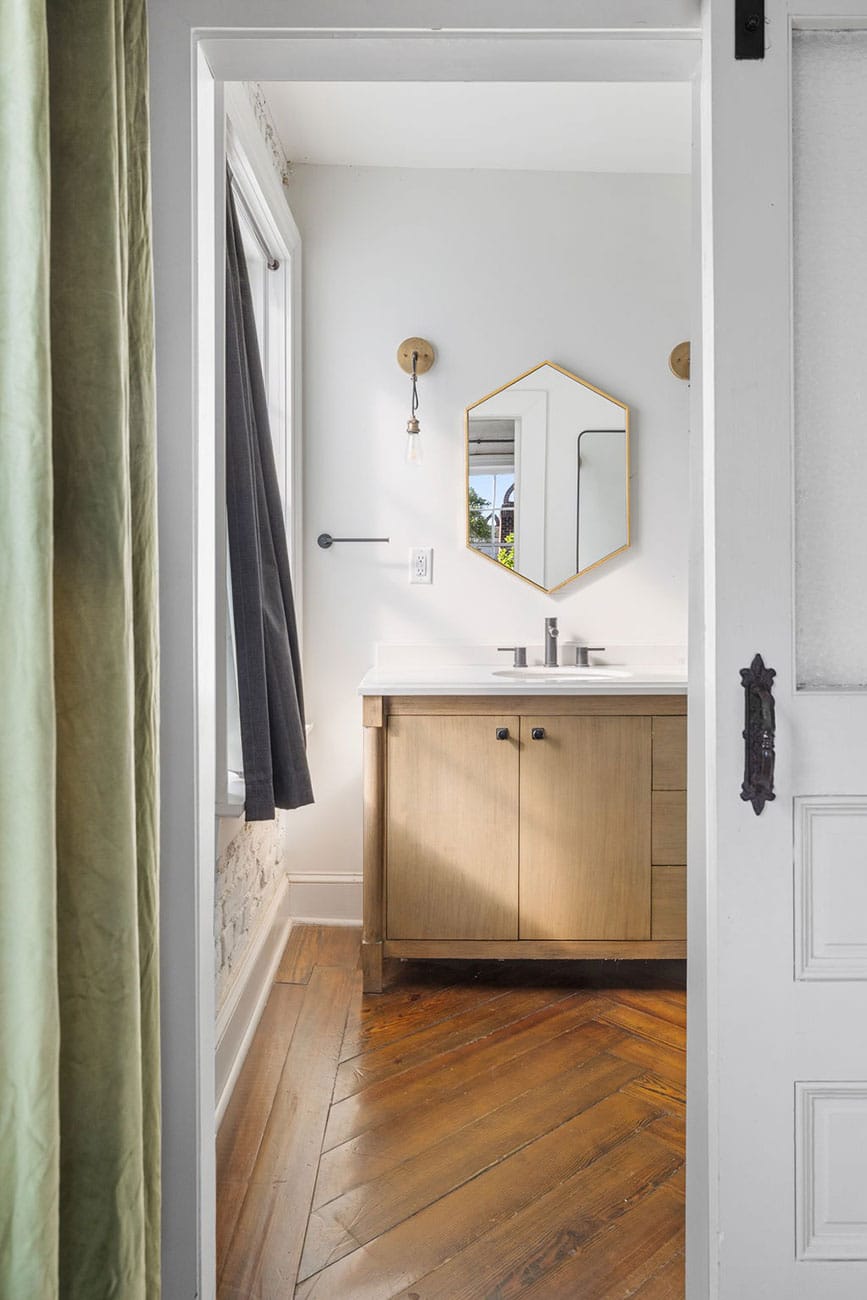 View into a small bathroom with a wooden vanity, hexagonal mirror, and warm wood floor