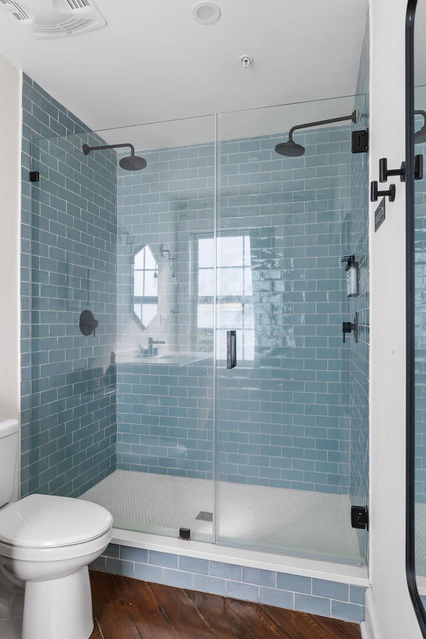 Modern glass-enclosed shower with blue subway tiles, two ceiling rain showers, and a white toilet nearby, in a contemporary bathroom.