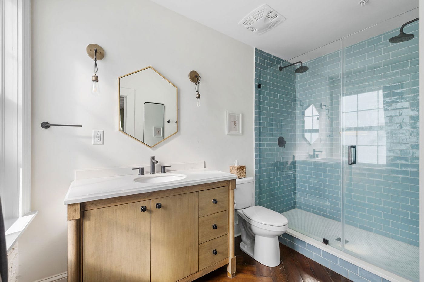 Bathroom with wooden vanity and white countertop, hexagonal mirror, and a glass-enclosed blue-tile shower area.