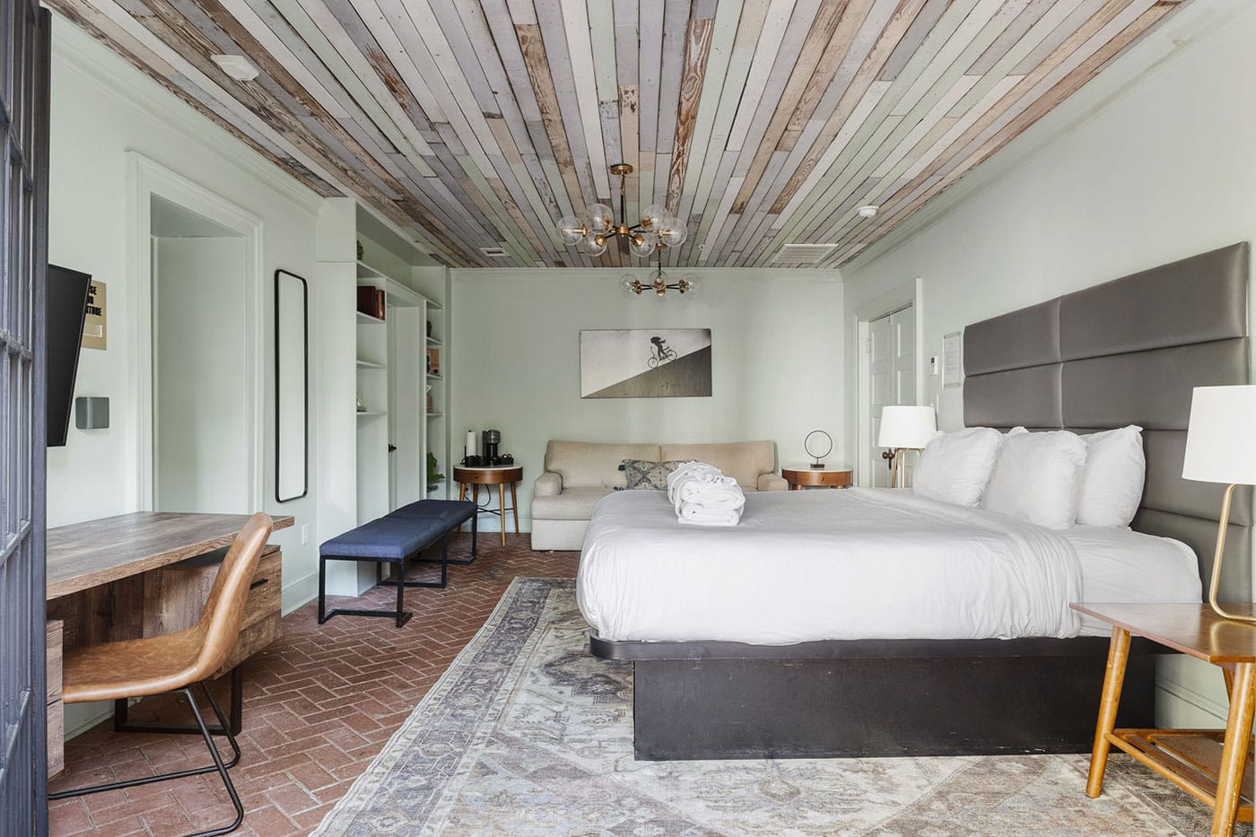 Hotel bedroom with a king bed, gray upholstered headboard, white linens, and a seating area under a wood-plank ceiling.