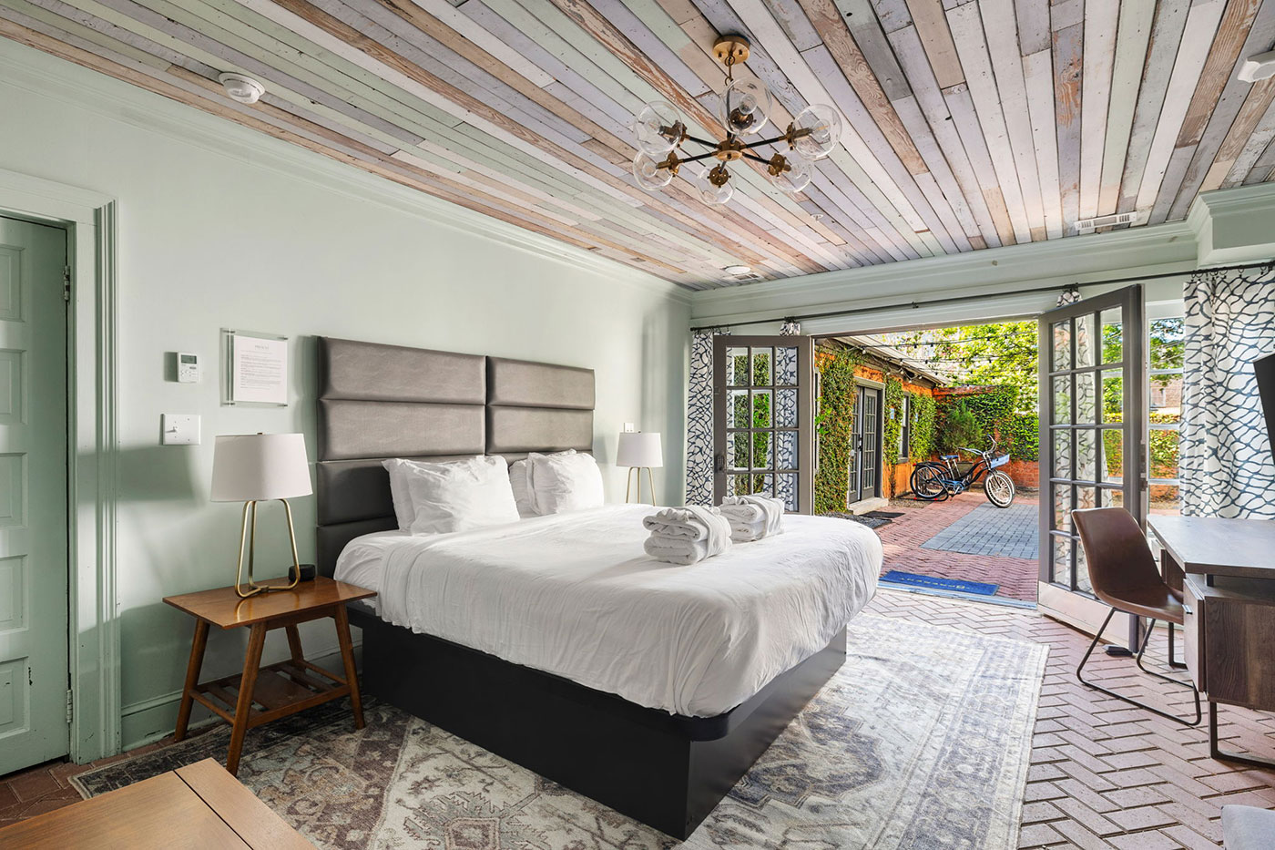 Bright hotel bedroom with a white bed, gray padded headboard, pastel green walls, and open French doors to a garden courtyard with a bicycle outside.