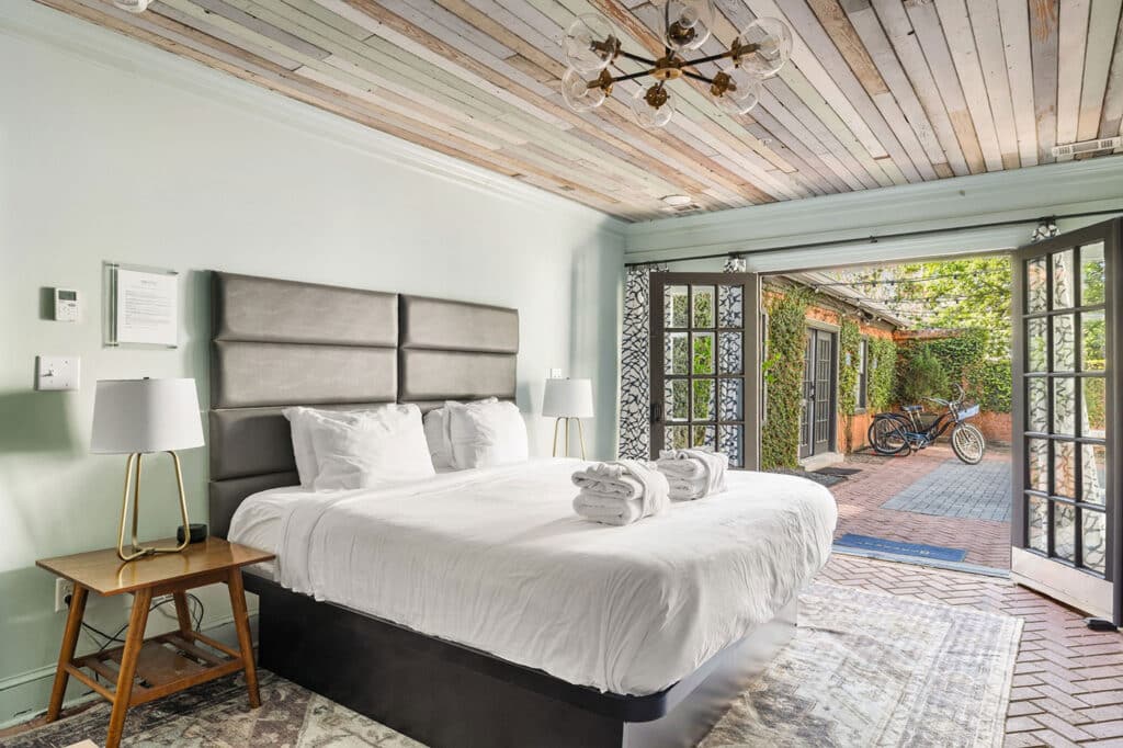 Bright hotel-style bedroom with a padded gray headboard, white bedding, and two bedside lamps, opening to a brick courtyard with a bicycle outdoors and ivy.