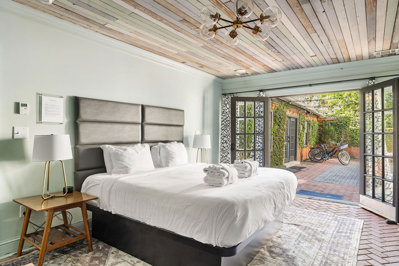 Bright hotel-style bedroom with a padded gray headboard, white bedding, and two bedside lamps, opening to a brick courtyard with a bicycle outdoors and ivy.
