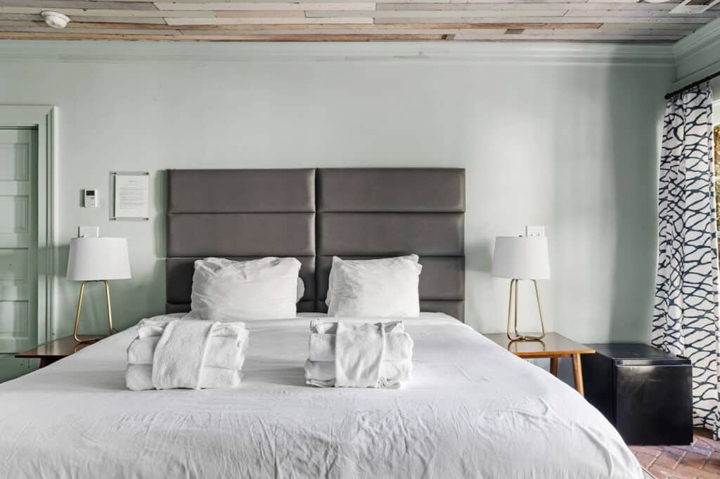 Hotel bedroom with a large bed, gray padded headboard, two white lamps on wooden nightstands, and rolled towels on the white duvet.