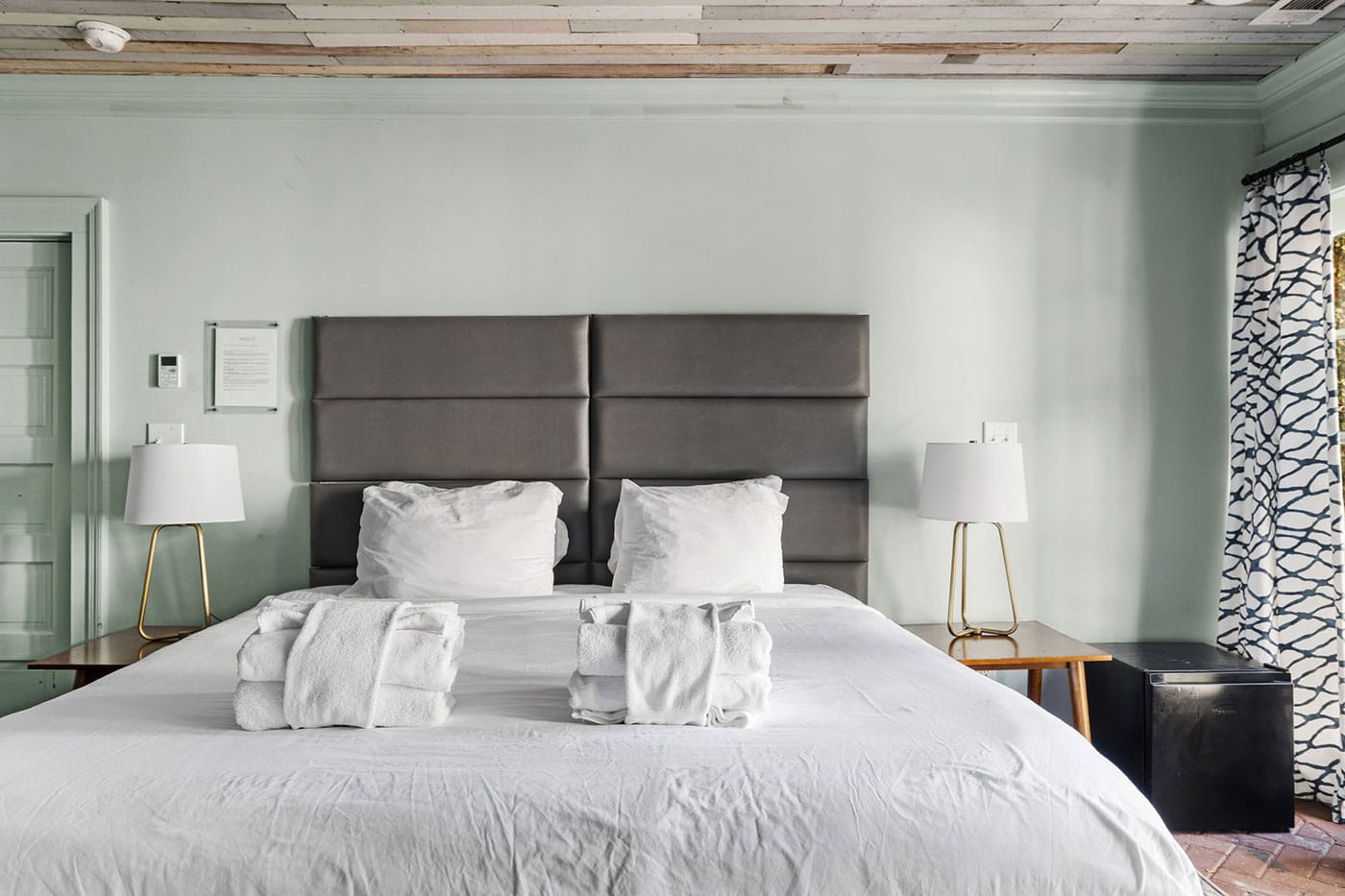Hotel bedroom with a large bed, gray padded headboard, two white lamps on wooden nightstands, and rolled towels on the white duvet.