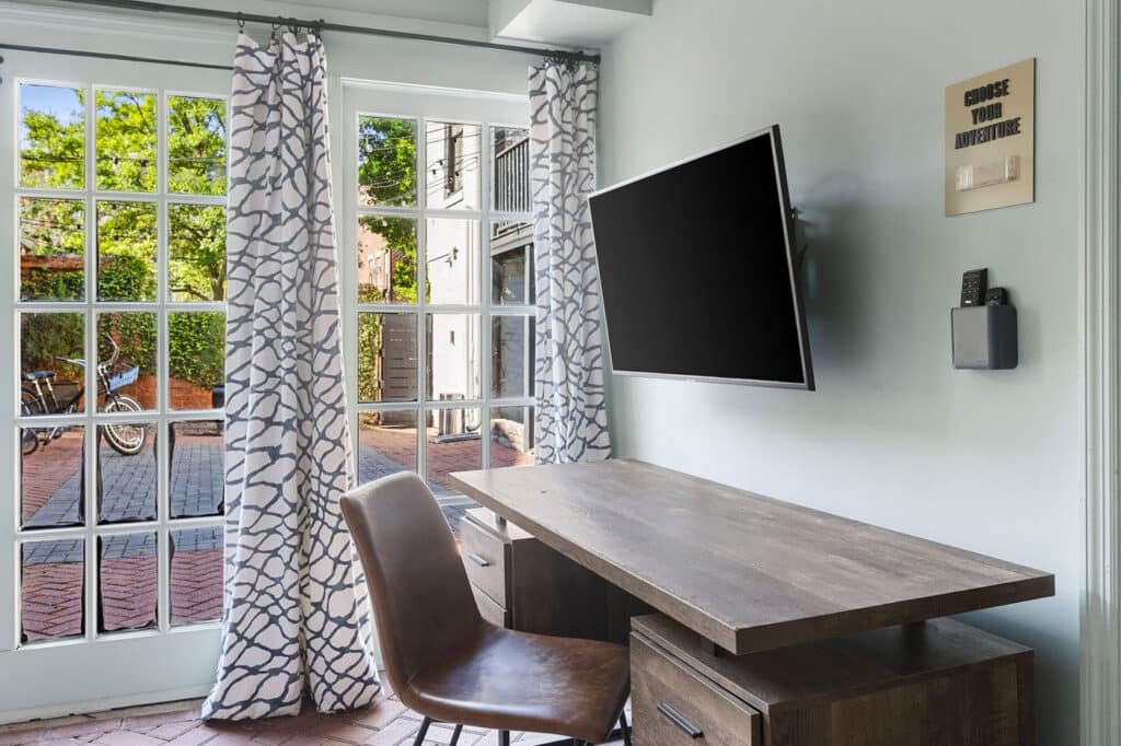 Modern home office with a wall-mounted flat-screen TV over a wooden desk and brown chair, sunny glass doors with patterned curtains reveal a courtyard outside.