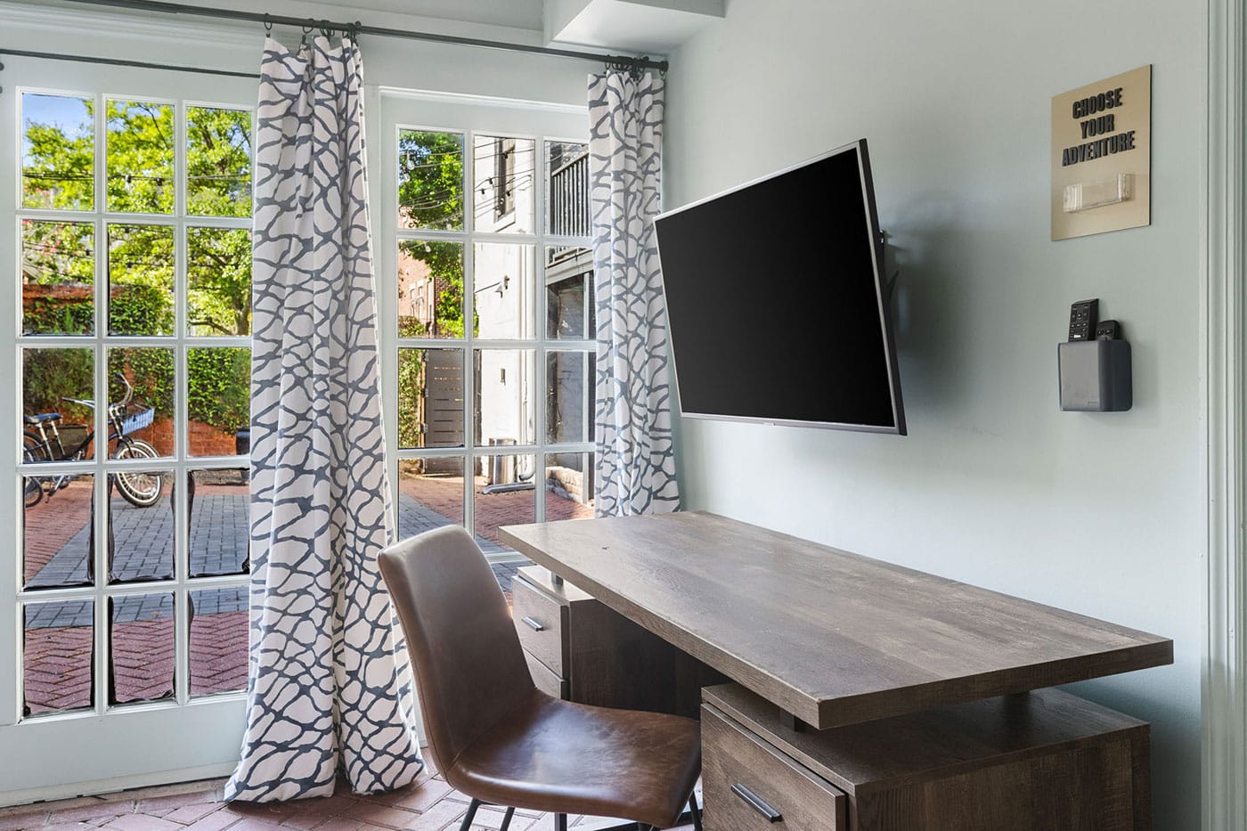 Modern home office with a wall-mounted flat-screen TV over a wooden desk and brown chair, sunny glass doors with patterned curtains reveal a courtyard outside.