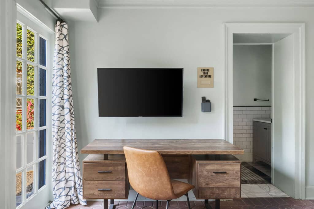 Wood desk with built-in drawers, a tan leather chair, and a wall-mounted flat-screen TV on a light green wall; window with patterned curtains to the left and a doorway to a tiled bathroom on the right.