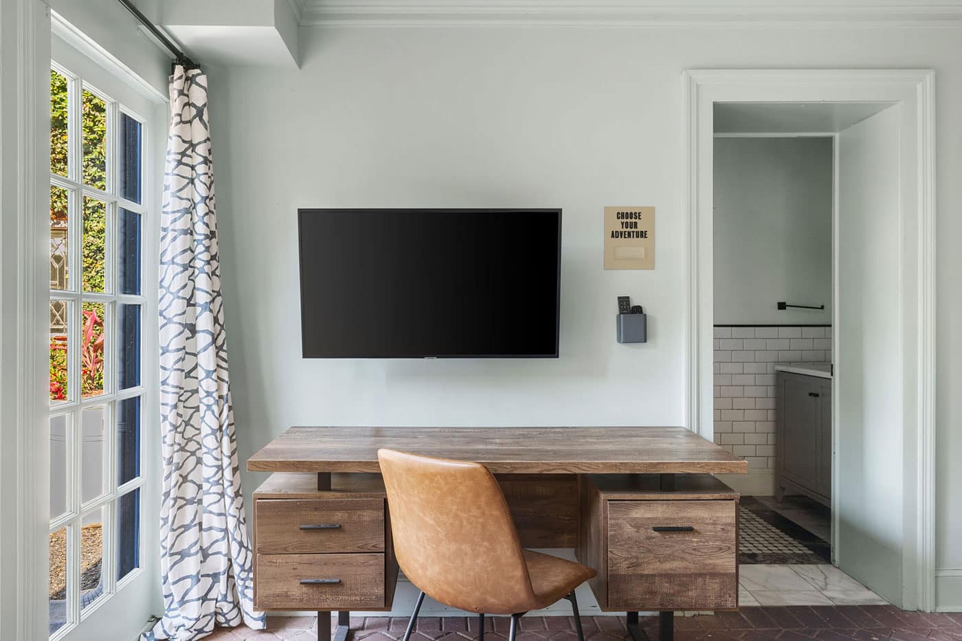 Wood desk with built-in drawers, a tan leather chair, and a wall-mounted flat-screen TV on a light green wall; window with patterned curtains to the left and a doorway to a tiled bathroom on the right.