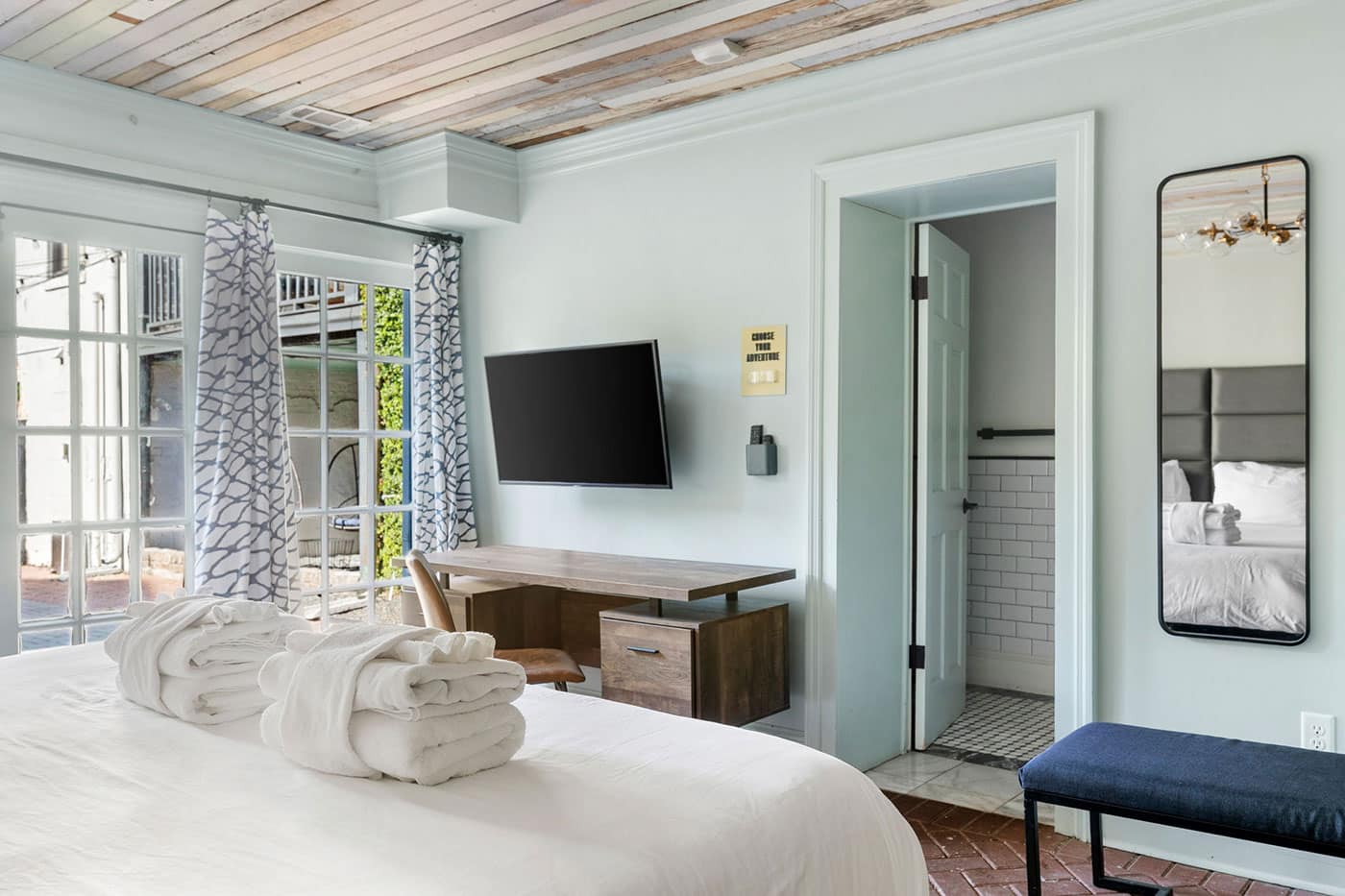 Hotel room with a large bed, folded white robes, a wall-mounted TV, and a wooden desk near French doors with patterned curtains; doorway to a tiled bathroom visible in the background.