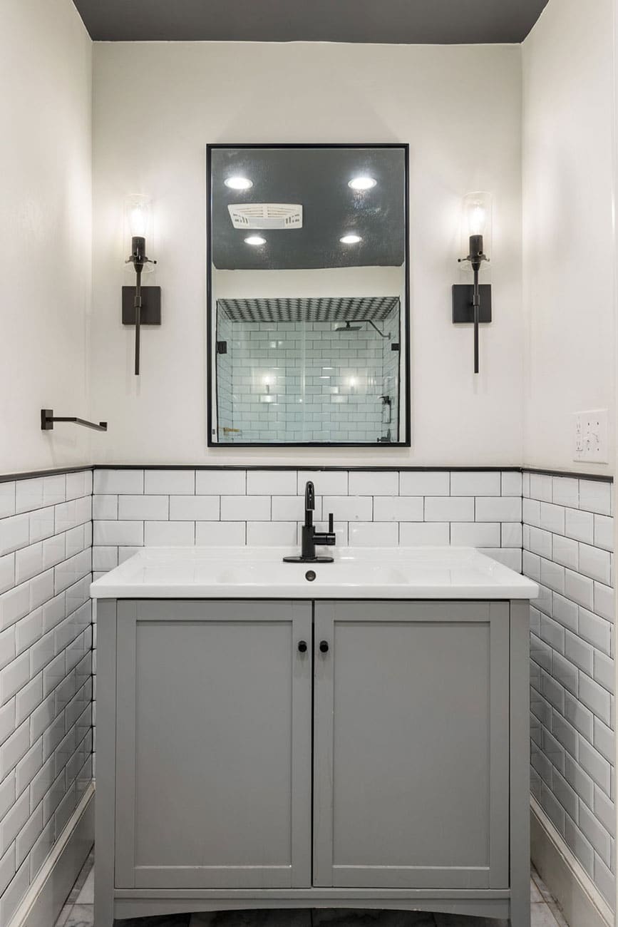 Gray bathroom vanity with white countertop and black faucet, framed mirror, and wall sconces on either side.