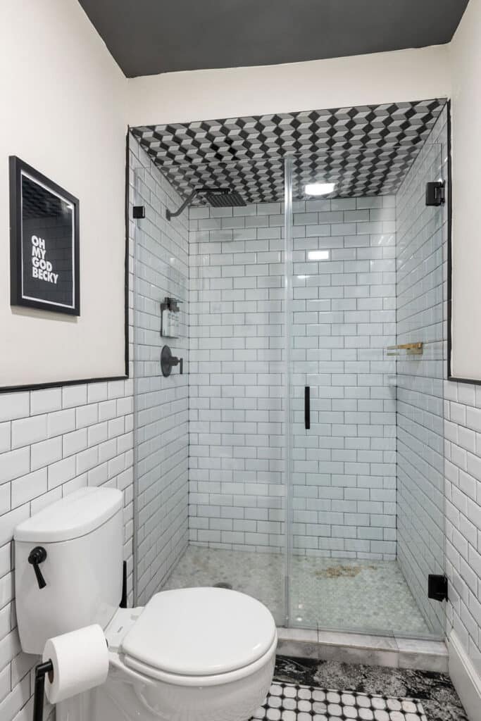 Framed print reading 'OH MY GOD BECKY' on a white-tiled wall beside a modern toilet in a bathroom with a glass-enclosed shower.