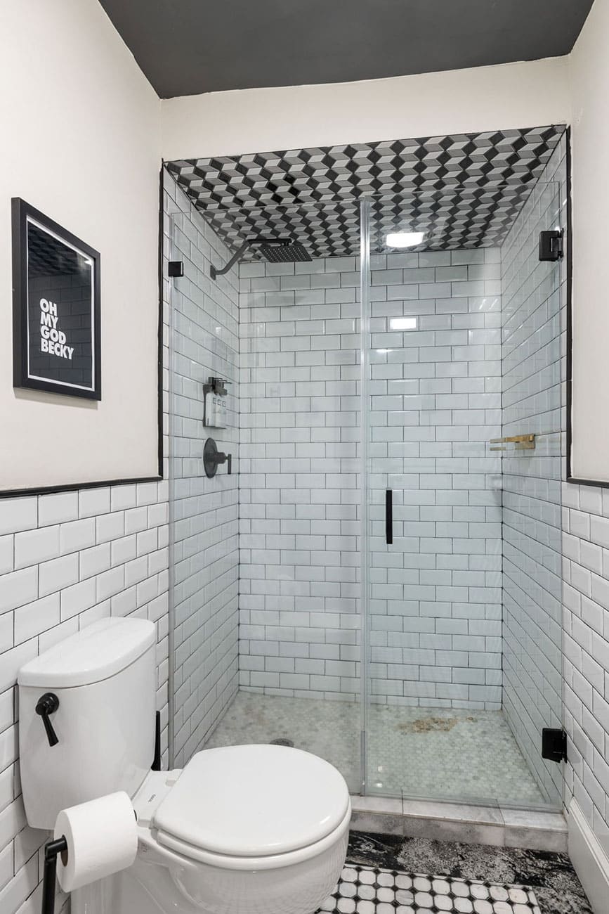 Framed print reading 'OH MY GOD BECKY' on a white-tiled wall beside a modern toilet in a bathroom with a glass-enclosed shower.
