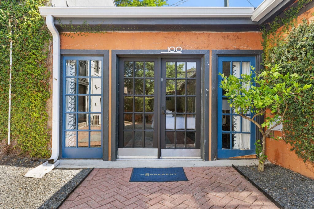 Blue-framed glass doors set in an orange building entrance, ivy on both sides, with a brick-paved path and small tree nearby.