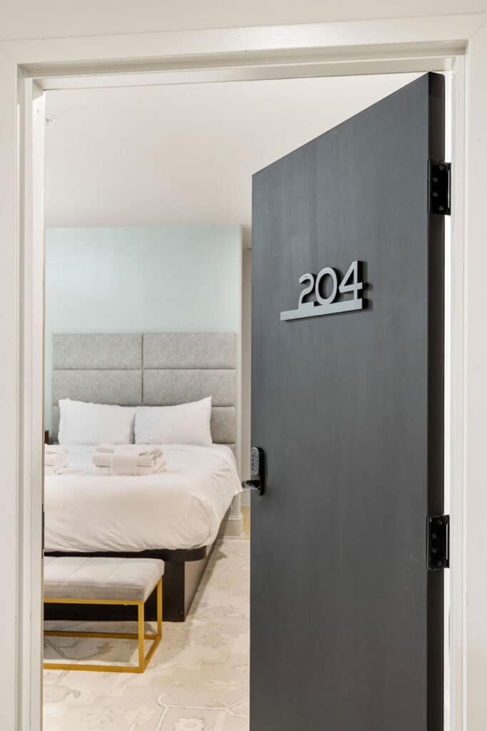 Door slightly ajar showing room number 204 and a hotel bedroom with a white bed, plush headboard, two pillows, folded towels on the bed, and a small bench at the foot of the bed.