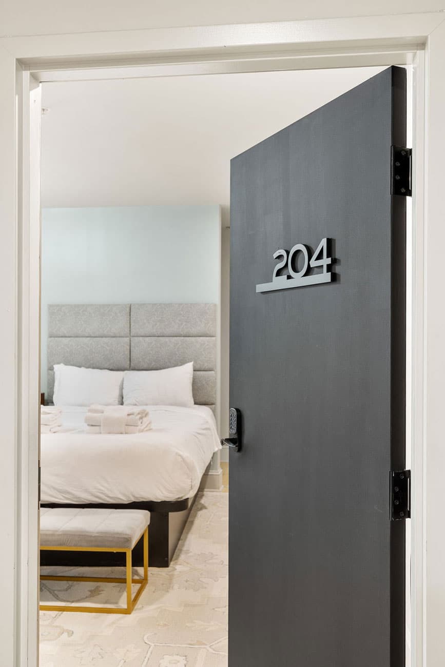 Door slightly ajar showing room number 204 and a hotel bedroom with a white bed, plush headboard, two pillows, folded towels on the bed, and a small bench at the foot of the bed.