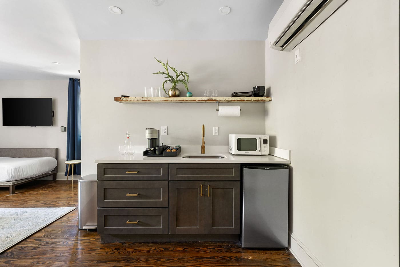 Studio kitchenette with dark wood cabinets, mini fridge, microwave, coffee maker, and a wooden shelf with plants above the sink area.