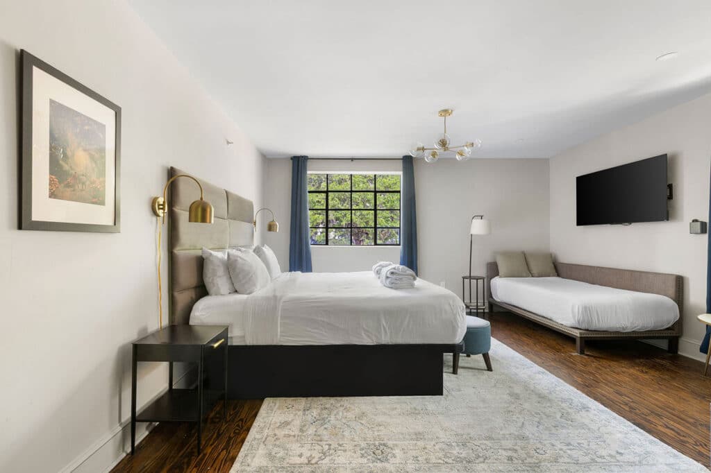 Hotel room with a large king bed, gray upholstered headboard, and gold wall lamps flanking the bed; window with blue curtains in the background.