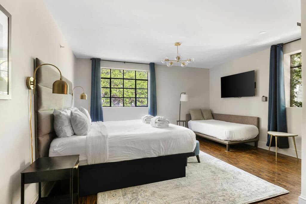 Spacious hotel bedroom with a king bed, gold curved wall lamps, an upholstered headboard, a second bed, blue curtains, and a wall-mounted TV. Visual balance of light and neutral tones inside a bright room.