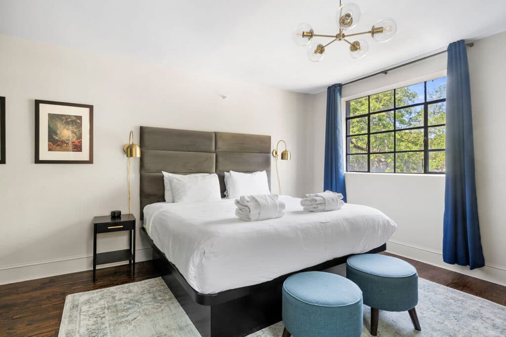 Bright hotel-style bedroom with a king bed, gray upholstered headboard, white linens, and two rolled towels on the bed, plus gold wall lamps on each side.