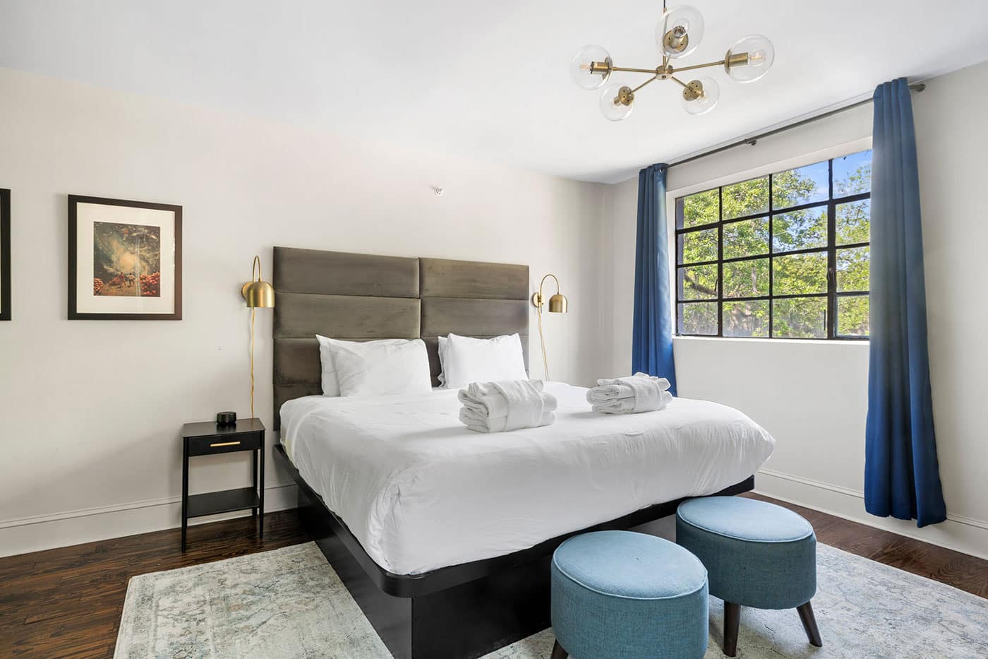 Bright hotel-style bedroom with a king bed, gray upholstered headboard, white linens, and two rolled towels on the bed, plus gold wall lamps on each side.