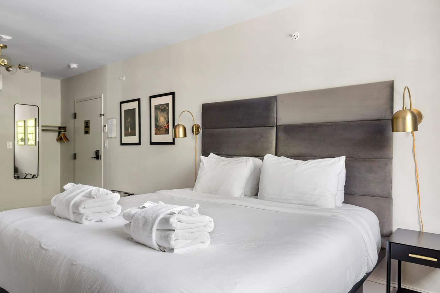 Hotel room with a large white bed, padded gray headboard, and two gold wall lamps; folded white robes sit on the bed.