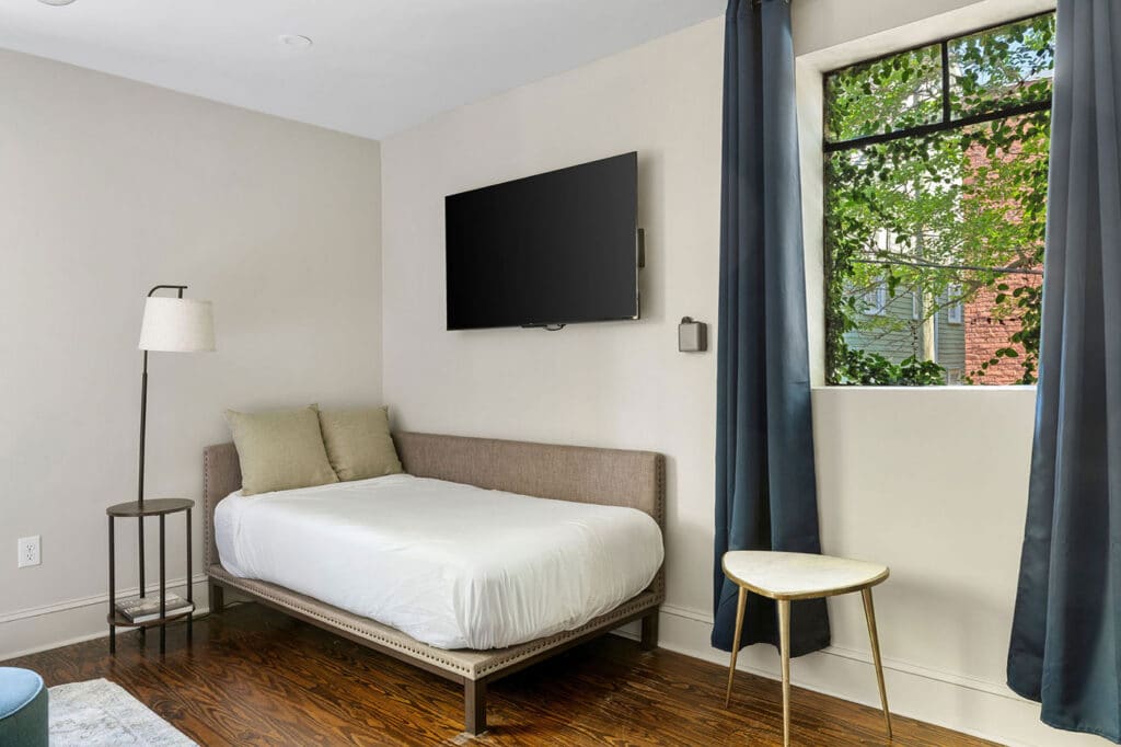 Bedroom with a single upholstered bed, two beige pillows, and a wall-mounted flat-screen TV above.