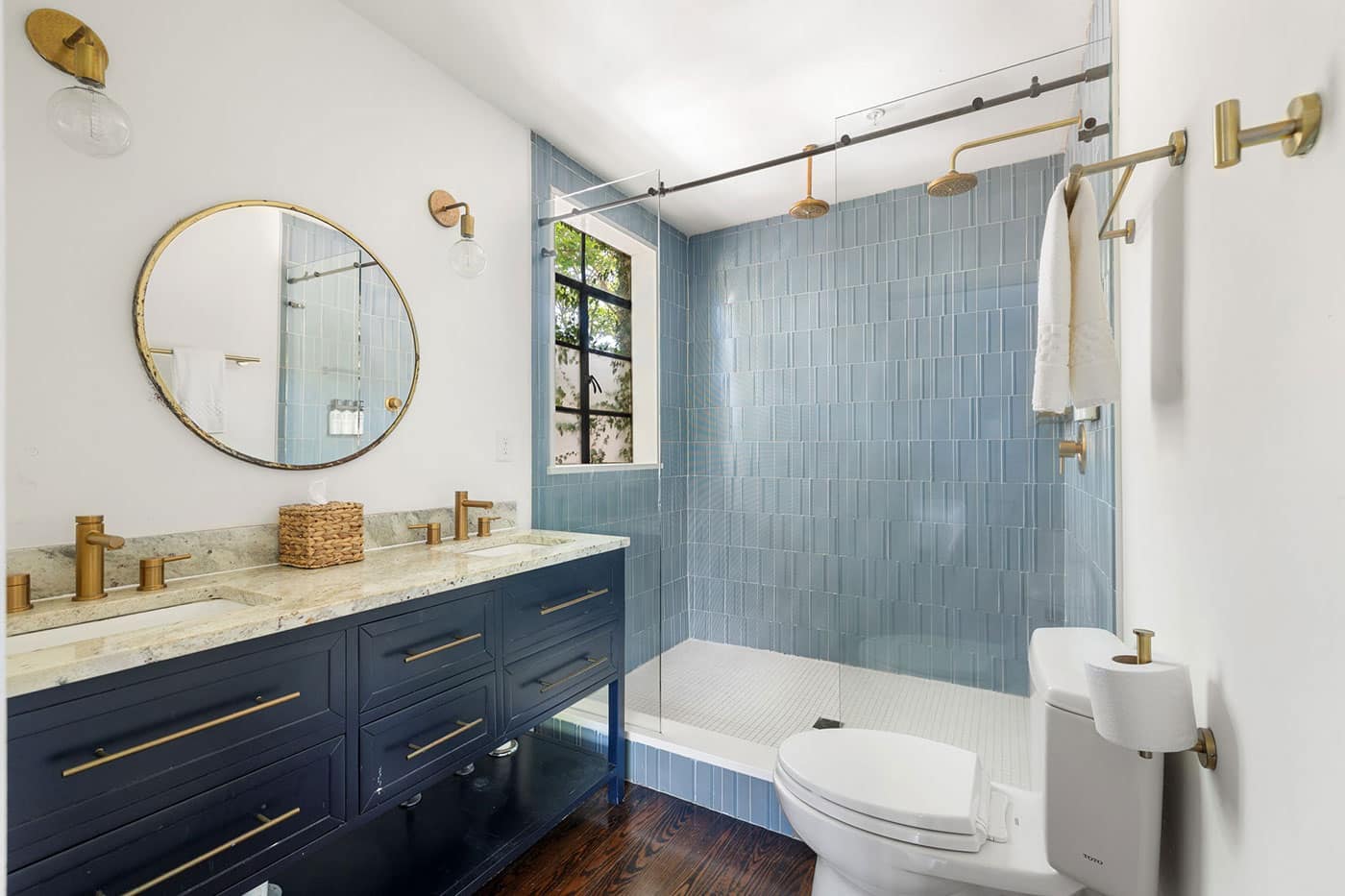 Modern bathroom with a double vanity in navy cabinets, marble countertop, brass faucets, and a round mirror reflecting a blue-tiled shower area with a glass enclosure.