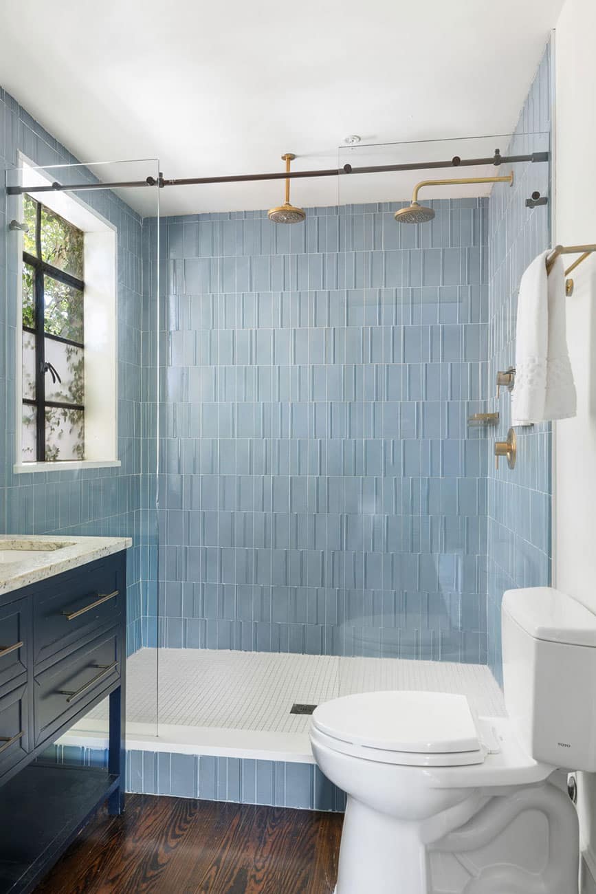 Blue tiled bathroom with a glass shower enclosure, two brass rainfall showerheads, a dark vanity, and a white toilet.