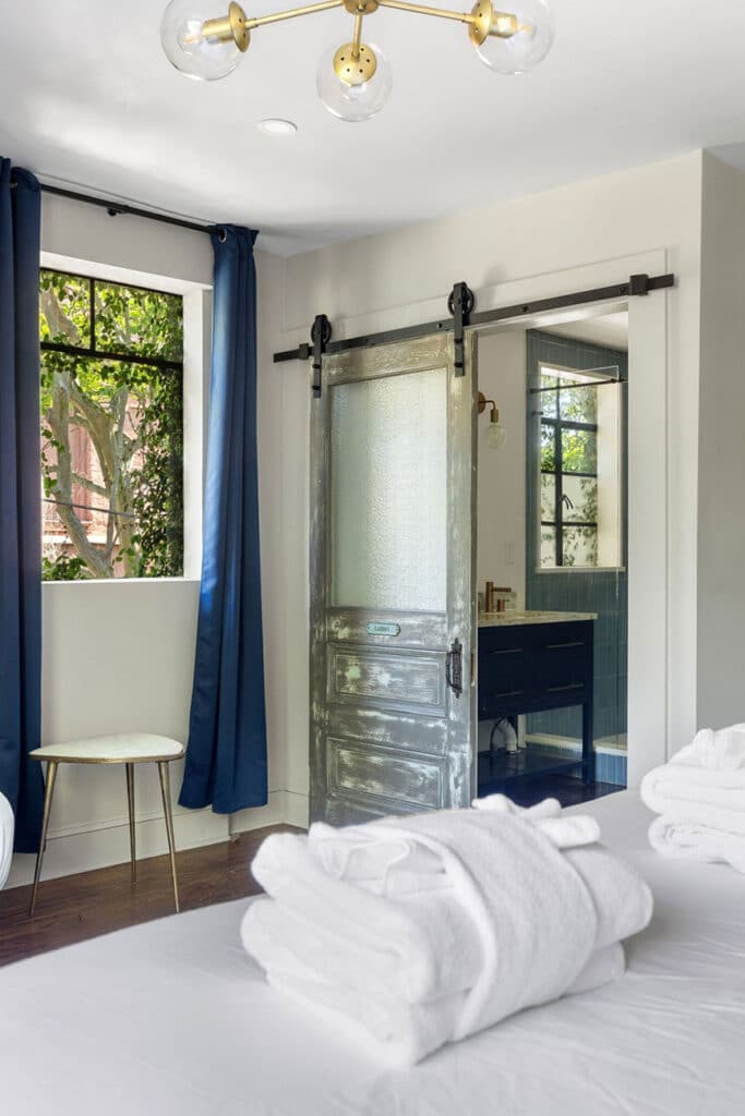 Bedroom with a frosted sliding barn door opening to a blue-tiled bathroom; navy curtains, a round white stool by the window, and folded white towels on the bed in the foreground.