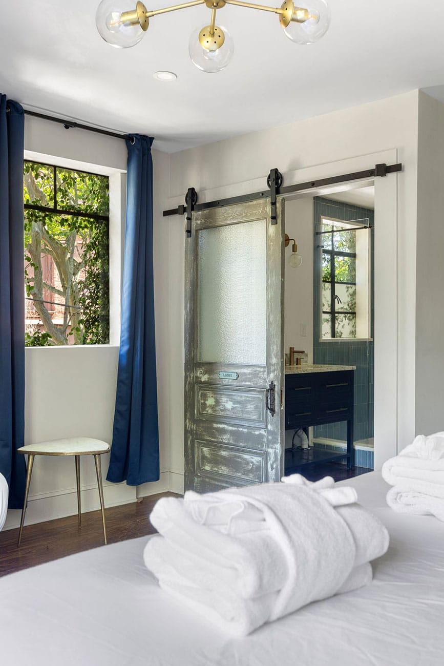 Bedroom with a frosted sliding barn door opening to a blue-tiled bathroom; navy curtains, a round white stool by the window, and folded white towels on the bed in the foreground.