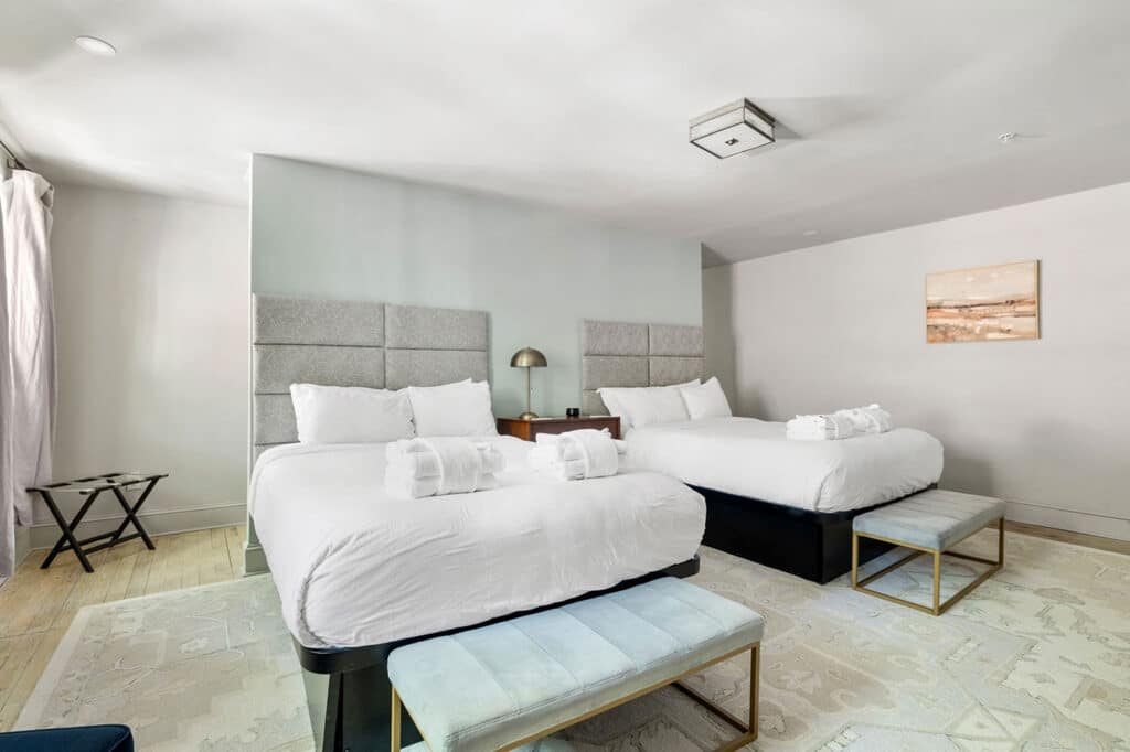 Hotel room with two white-duvet beds, padded gray headboards, a nightstand with a brass lamp between them, and rolled towels on each bed.