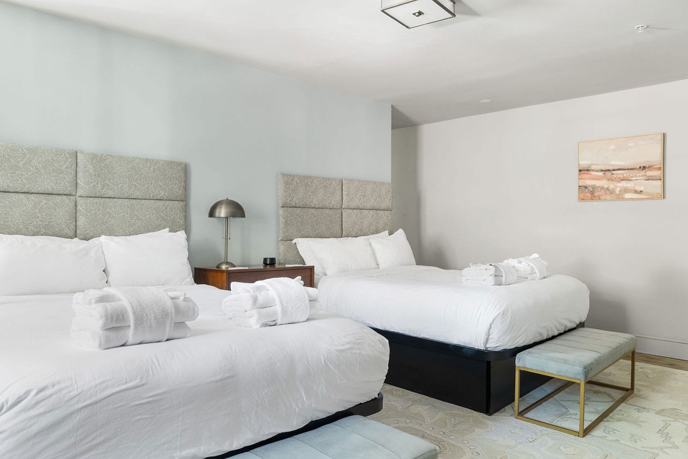 Hotel room with two large beds in white linens, rolled towels on each bed, and a nightstand with a lamp between them.