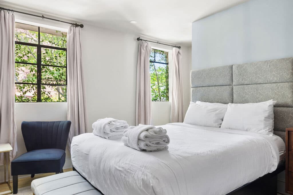 Bright bedroom with a white bed and gray padded headboard, two folded towels on the duvet, and a navy chair by a window.