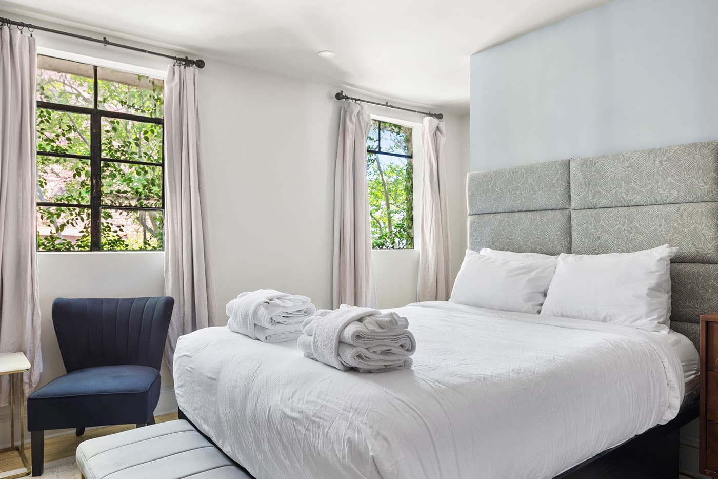 Bright bedroom with a white bed and gray padded headboard, two folded towels on the duvet, and a navy chair by a window.