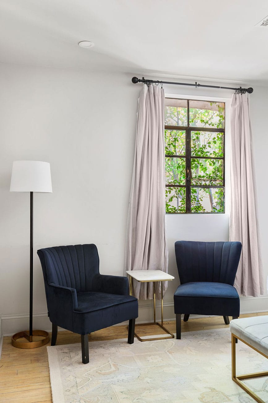 Two navy upholstered armchairs flank a small marble-top side table in a bright living room with a floor lamp and a window with light pink curtains.