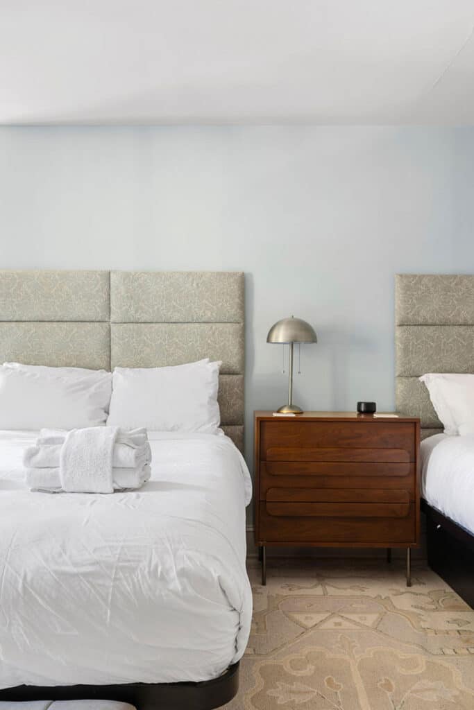 Hotel bedroom with two single beds, white linens, folded towels on the left bed, and a wooden nightstand with a lamp between them.