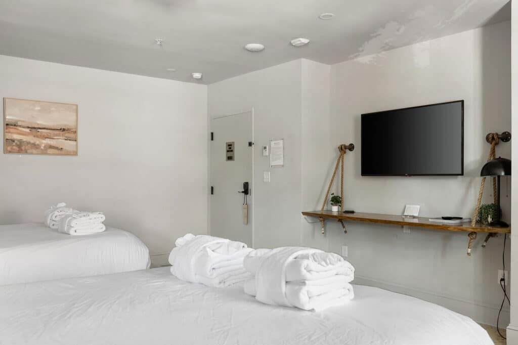 Hotel room with two white beds, neatly folded towels, and a wall-mounted flat-screen TV on a rope-supported shelf.